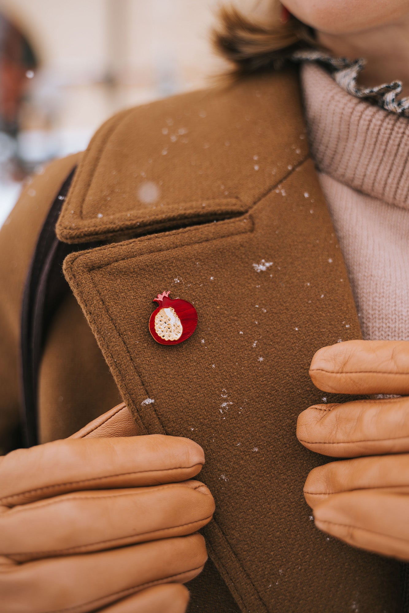 A person wearing a brown coat and tan gloves adjusts the lapel, which is decorated with a handcrafted Pomegranate Pin—a unique fashion accessory. Snowflakes are visible on the coat.
