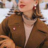 A stylish woman in a beige beret, brown coat, and tan gloves poses outdoors in snowy weather. She wears red and gold handcrafted earrings and a matching Pomegranate Pin fashion accessory, with snowflakes visible on her coat.
