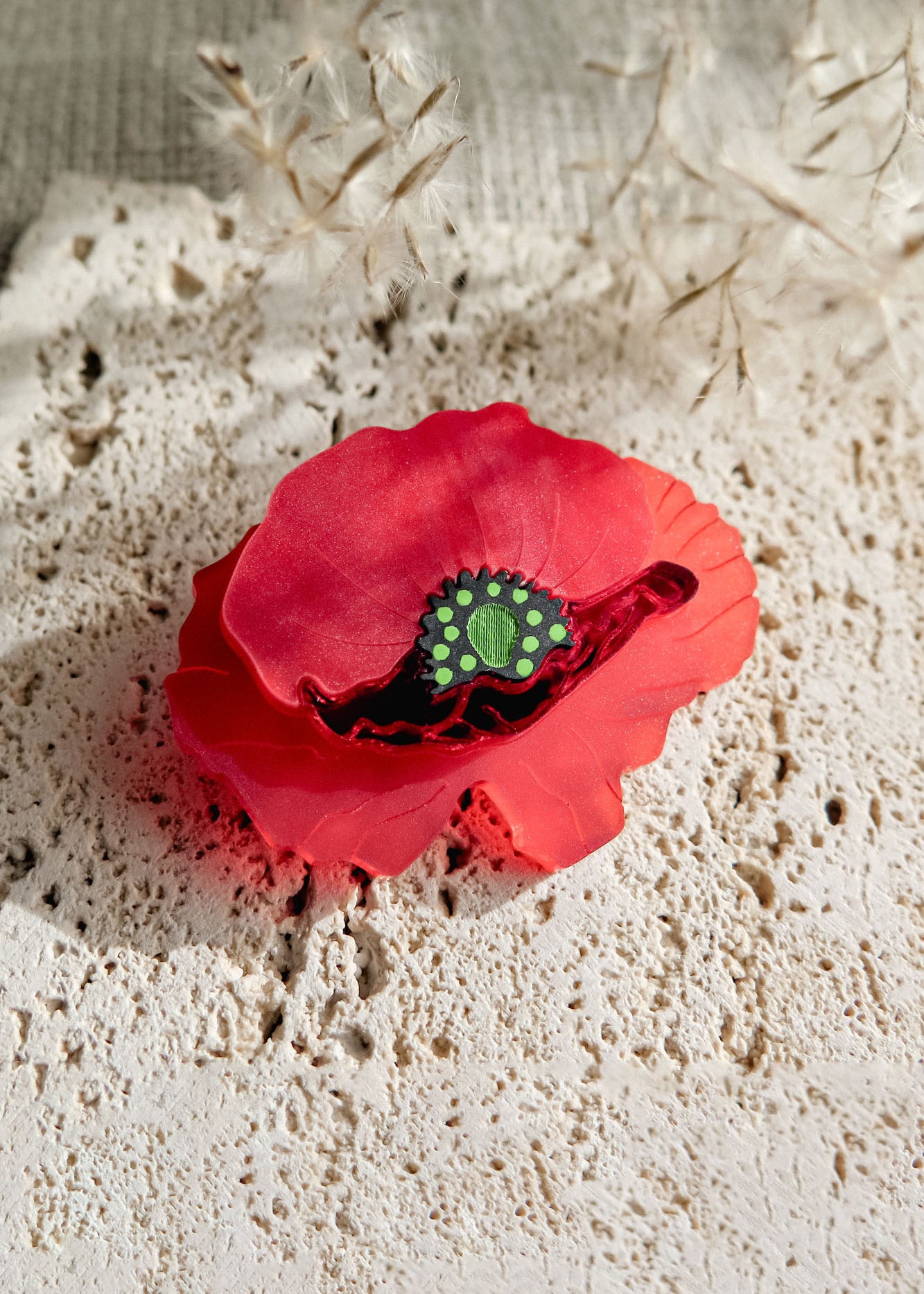 A red brooch clip shaped like a flower with a green acrylic glass center rests on a textured, off-white stone surface, accented by delicate dried plants in the background.