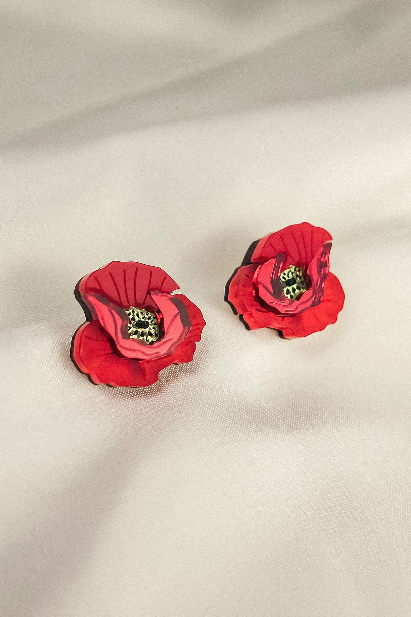 A pair of red, poppy design acrylic earrings with black and green centers, displayed on a soft, light-colored fabric background. Lightweight and eye-catching for any occasion.