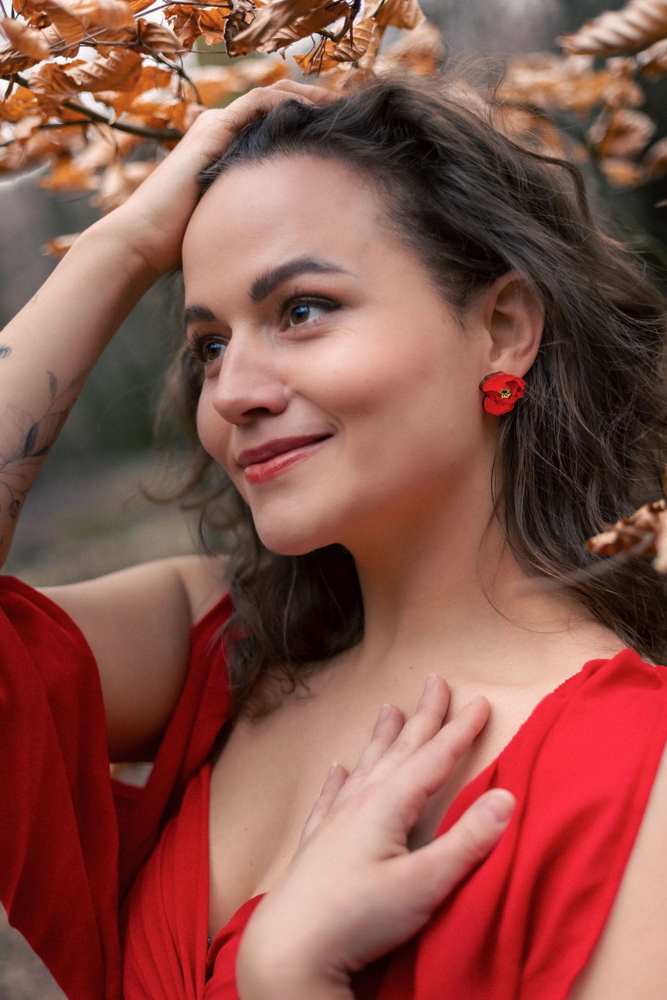 A woman with wavy brown hair, wearing a red dress and lightweight acrylic earrings with a poppy design, stands outdoors among autumn leaves, smiling softly with one hand on her head and the other on her chest.