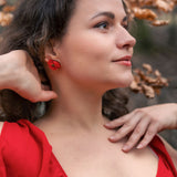 A woman in a red dress poses outdoors near branches with dried leaves. She touches her neck and head, looking to the side with a calm expression. Her wavy brown hair frames lightweight acrylic earrings and a striking red earring.