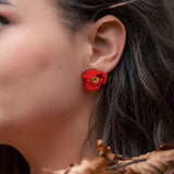Close-up of a womans ear adorned with a bright red poppy design acrylic earring. Part of her face and dark, wavy hair are visible, with dried brown leaves in the foreground. The earring is lightweight and eye-catching.