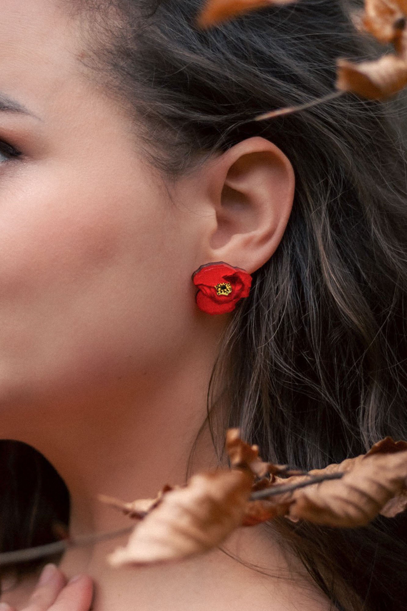 Close-up of a womans ear adorned with a bright red poppy design acrylic earring. Part of her face and dark, wavy hair are visible, with dried brown leaves in the foreground. The earring is lightweight and eye-catching.