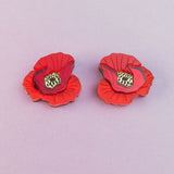 Two lightweight acrylic earrings featuring a vibrant poppy design with black and yellow centers are displayed on a light purple background.