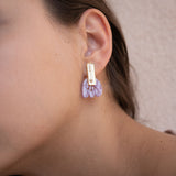 Close-up of a person wearing a gold rectangular earring with marbled purple acrylic petal-shaped accents, against a light beige background. Showcased on the left ear, these Romantic Tulip Earrings offer allergy-free comfort.