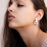 A woman with long brown hair gazes upward, showing off Romantic Tulip Earrings made from marbled purple acrylic. She wears a light pink, textured top, and the background is neutral for allergy-free comfort all day.