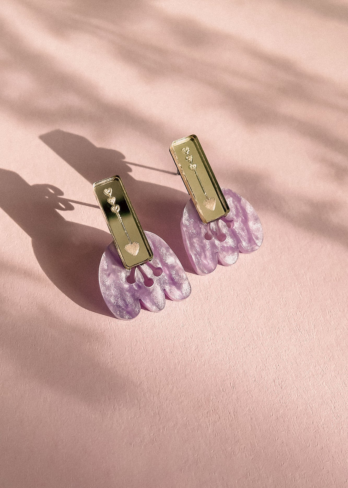 A pair of Romantic Tulip Earrings with rectangular gold tops and dangling, marbled purple acrylic flower-shaped pendants, displayed on a soft pink textured surface with delicate shadows for allergy-free comfort.