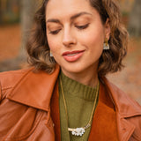 A woman with wavy brown hair, wearing a rust-colored jacket, olive green sweater, gold necklace with a floral pendant, and handcrafted Romantic Tulip Earrings, smiles gently with her eyes closed against an autumnal outdoor background.