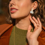 A woman with wavy, shoulder-length hair wears a green sweater, brown jacket, and gold handcrafted earrings—Romantic Tulip Earrings—with a matching necklace, gently touching her ear and smiling softly in warm sunlight.