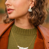 A woman with short wavy brown hair wears a green sweater, brown jacket, and an elegant Tulip Necklace with matching earrings featuring white accents, standing outdoors with blurred autumn foliage in the background.