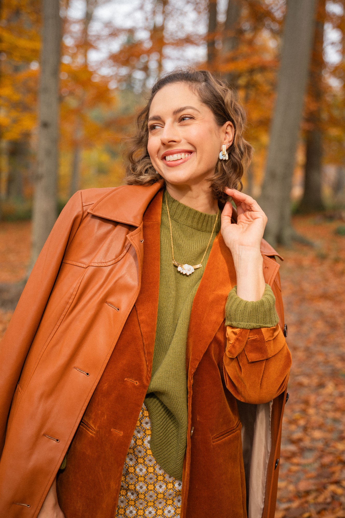 A woman smiles elegantly while standing in a forest with autumn leaves. She wears a brown leather coat over an orange suede jacket, with a green sweater, patterned skirt, and a handcrafted Tulip Necklace. Tall trees and fallen leaves fill the background.