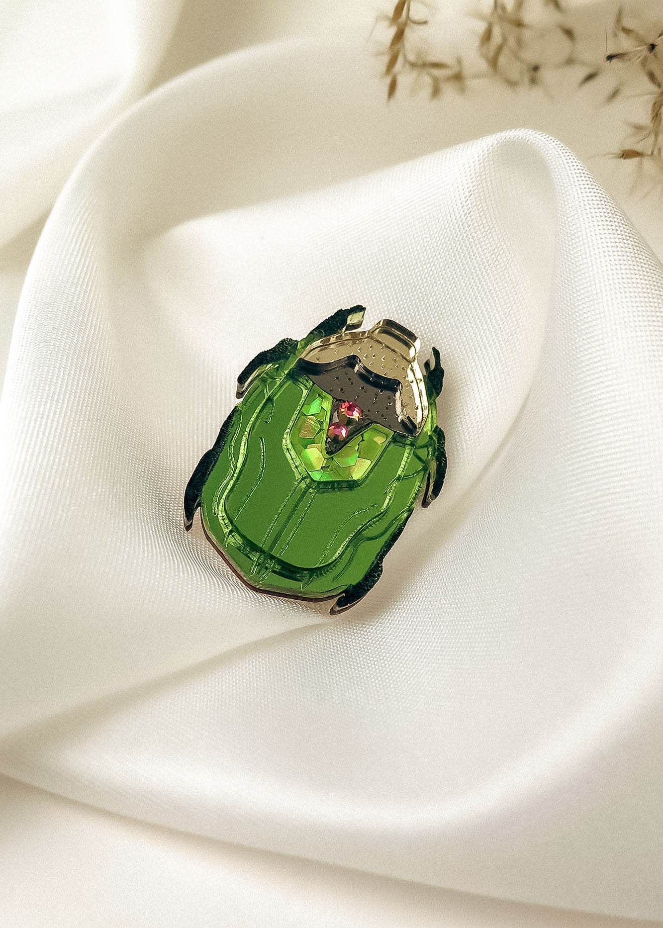 A handcrafted accessory, this green Rose Chafer Pin with black legs and silver accents rests on smooth white satin fabric, a few dried plant stems in the background highlighting its meaningful design.