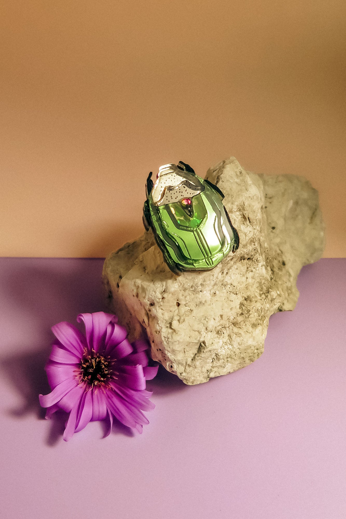 A green and white Rose Chafer Pin brooch with black accents rests on a light-colored rock next to a purple flower, all placed on a pastel purple surface with a beige background—an elegant, handcrafted accessory with meaningful design.