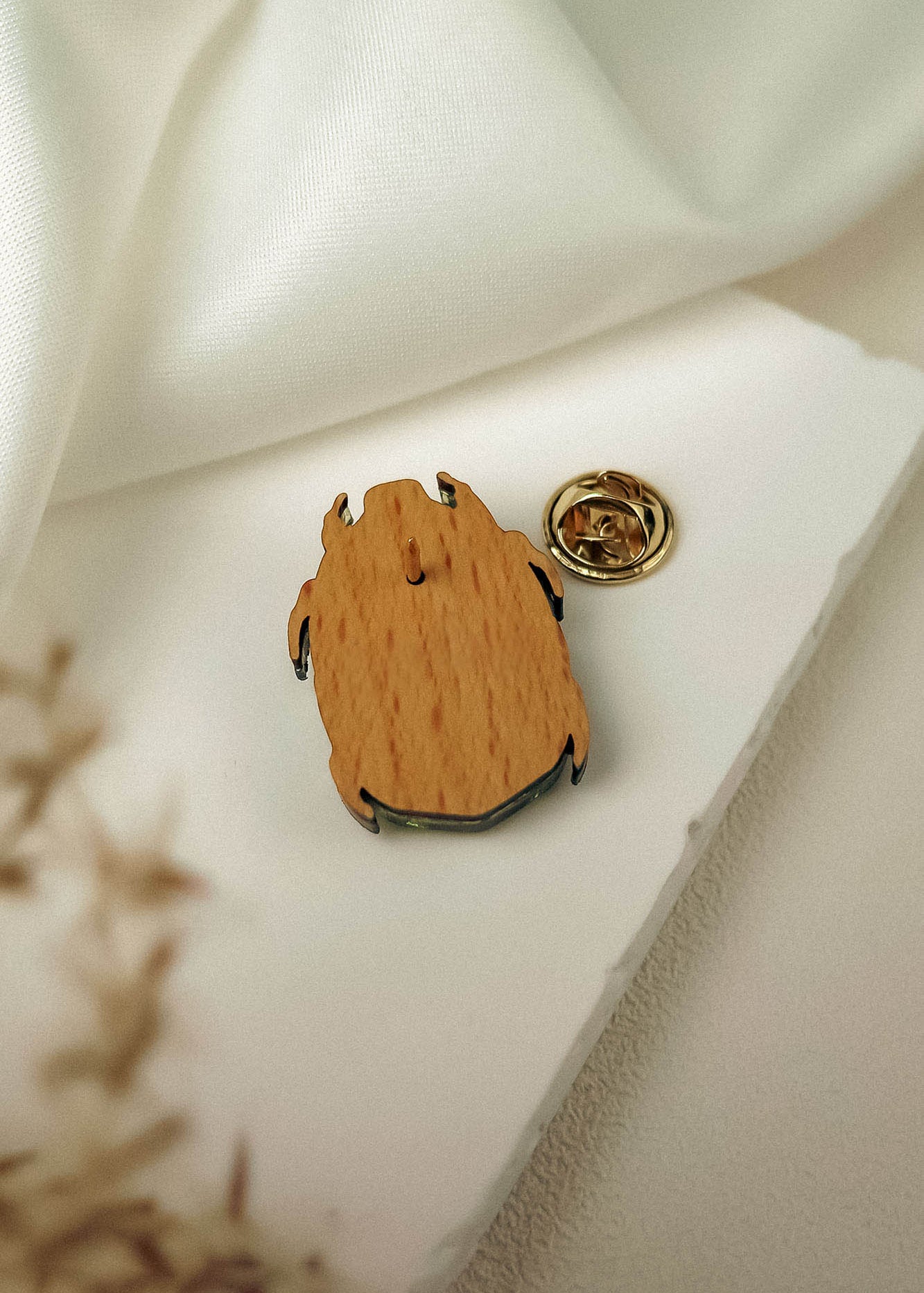 A wooden pin with a metal backing rests on a white surface near a piece of white fabric. This handcrafted accessory features an abstract, organic shape, and a gold-colored clasp is placed nearby.