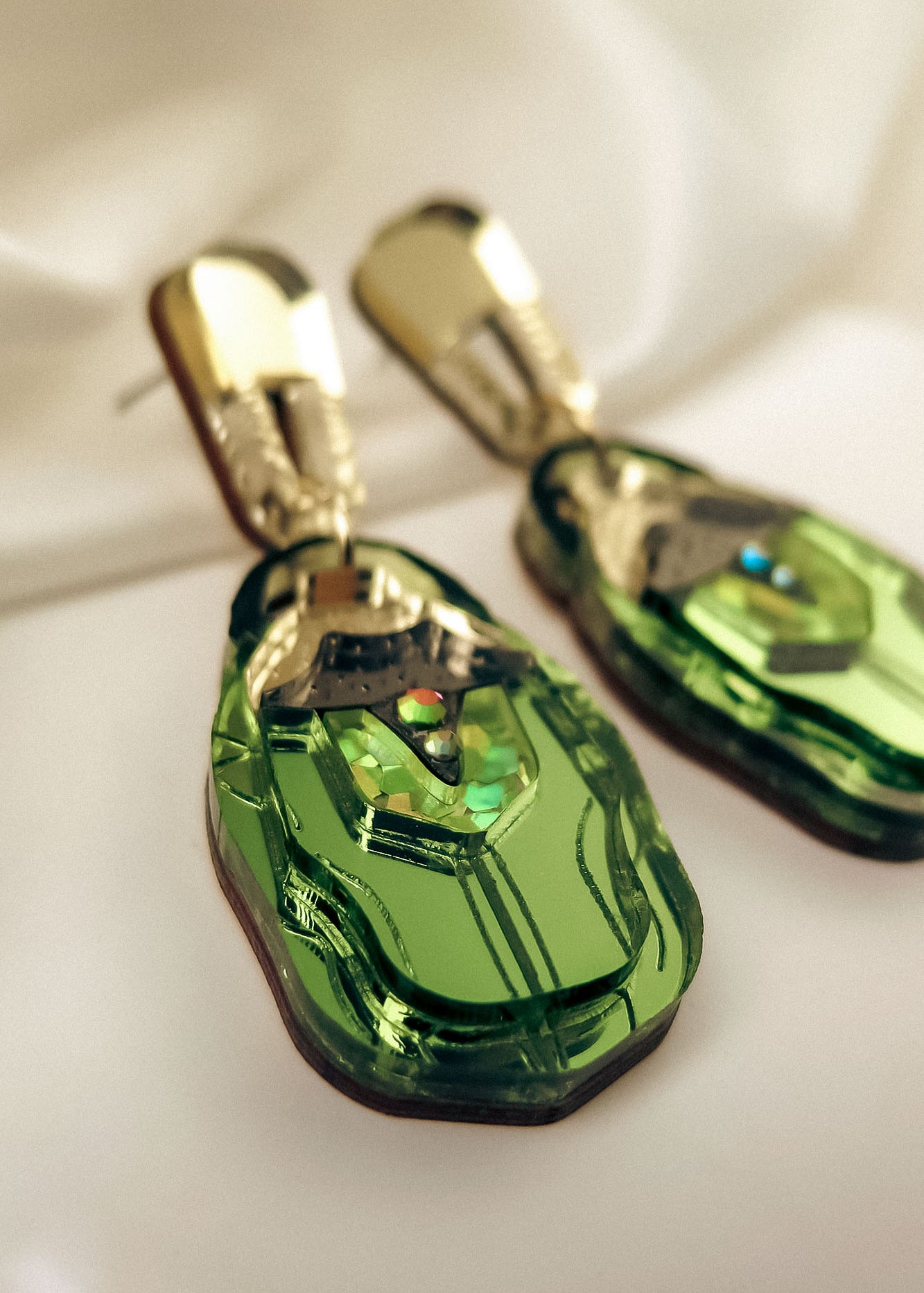 A close-up of handcrafted Rose Chafer Earrings, featuring gold and green geometric shapes with a shiny metallic finish, placed on a soft white fabric background for a nature-inspired look.