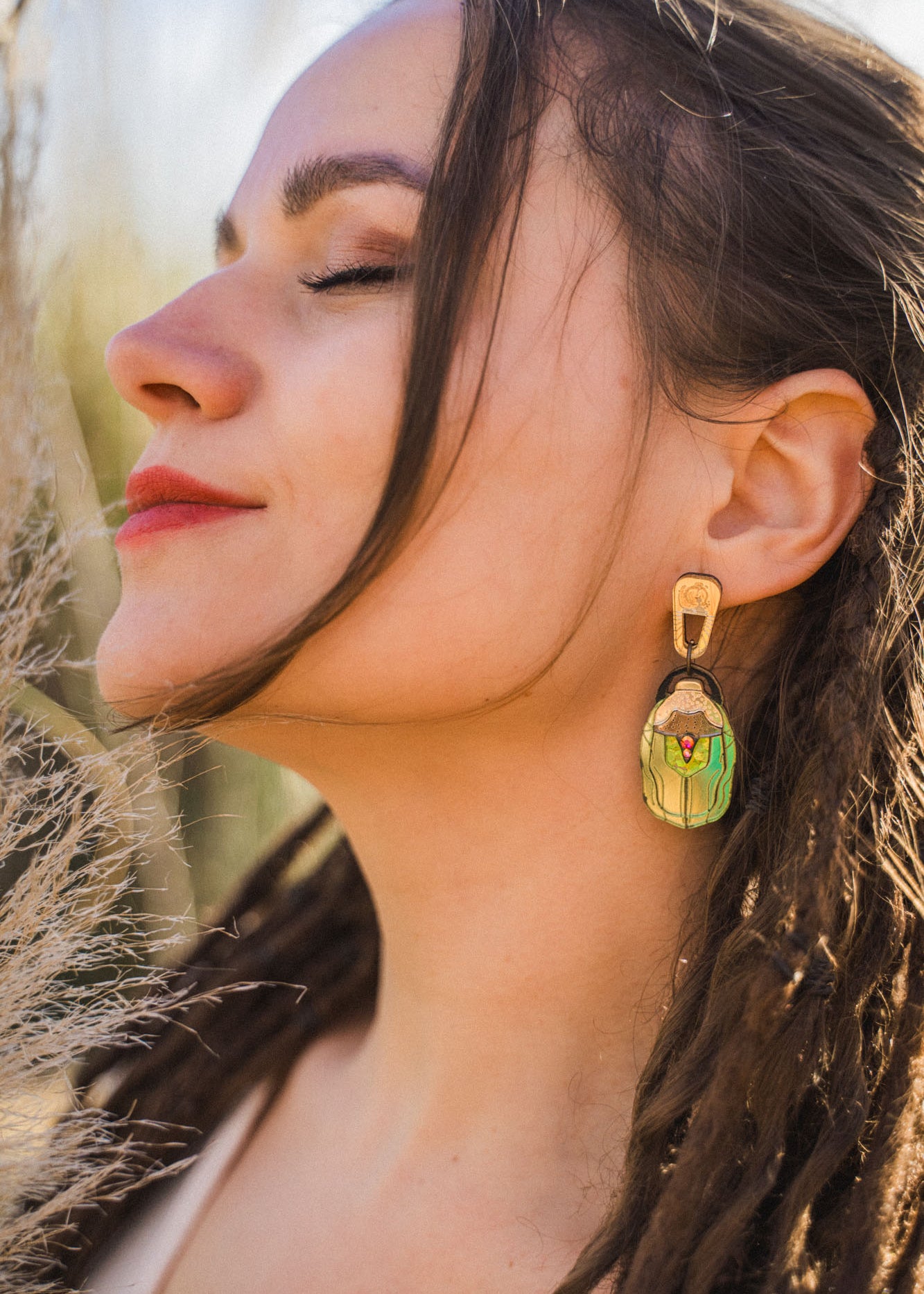 A woman with closed eyes and red lipstick faces the sunlight, wearing a large, nature-inspired earring shaped like a matryoshka doll. Her long brown hair is styled in loose dreadlocks.