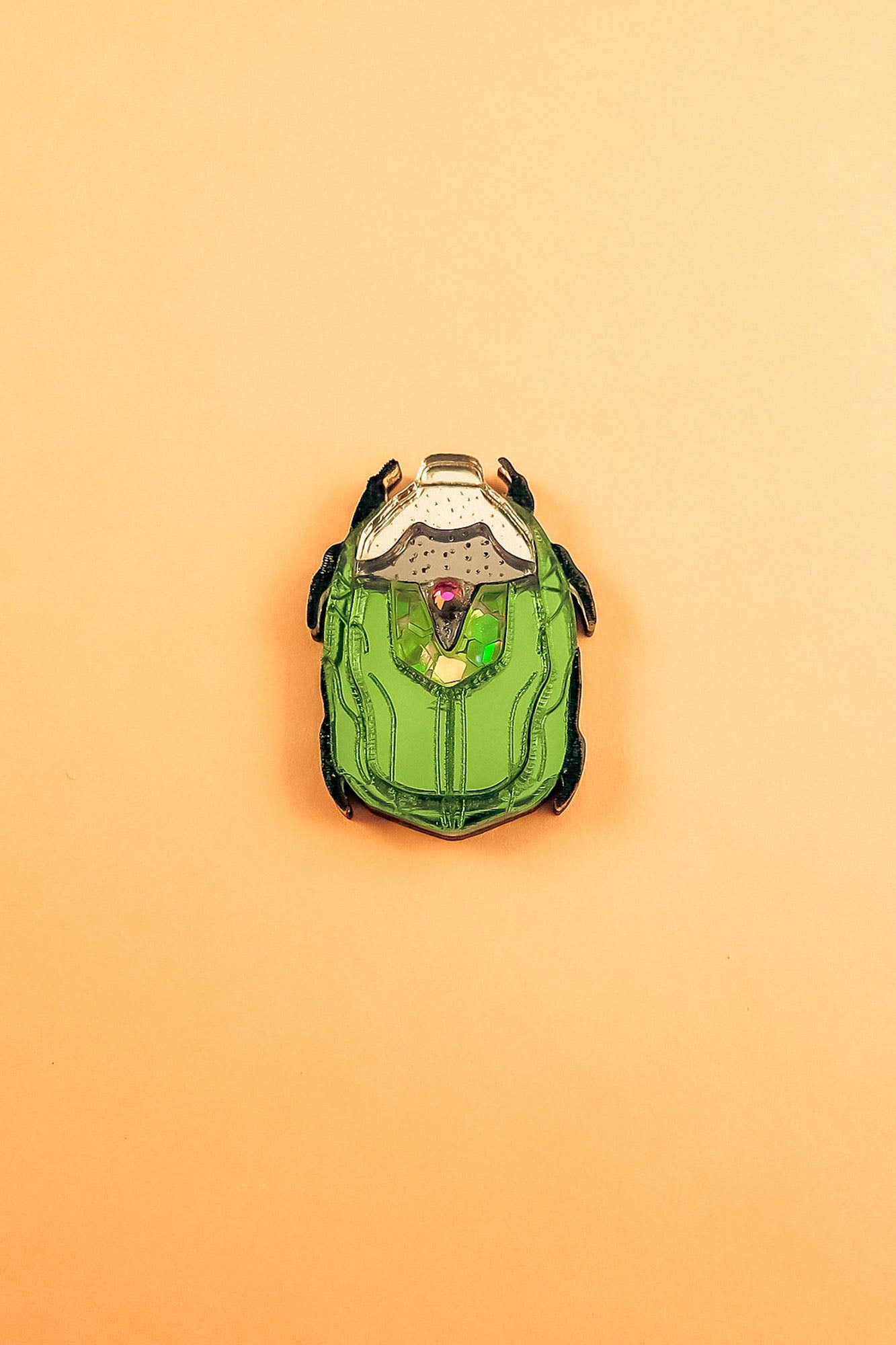 A green and black enamel Rose Chafer Pin, resembling a sci-fi or robot helmet, is centered on a plain peach background—a meaningful design perfect for lovers of handcrafted accessories.