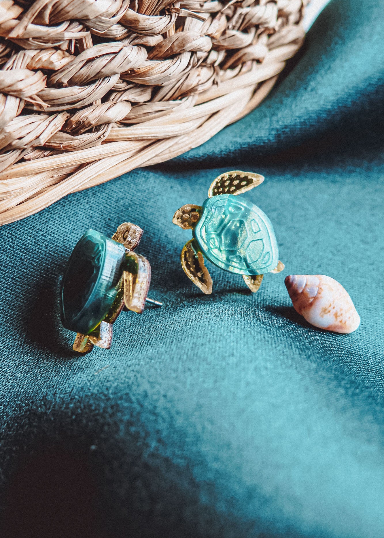 Two Sea Turtle Studs with green shells rest on a teal fabric beside a small seashell, evoking an ocean-inspired, summery vibe. A woven basket is visible in the background.