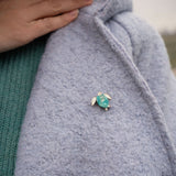 A person wearing a light blue textured coat with a handcrafted sea turtle pin in turquoise and silver on the lapel, and a green sweater underneath. Their hand is gently resting near the collar.