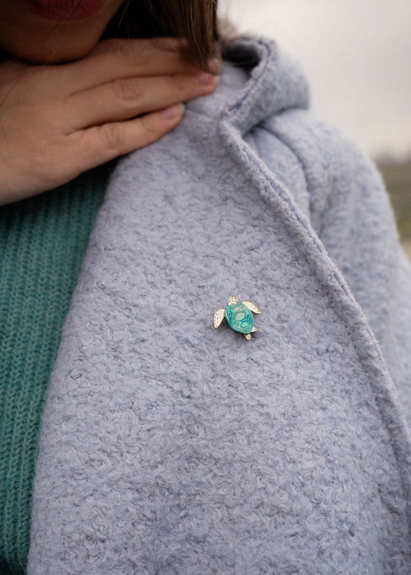 A person wearing a light blue textured coat with a handcrafted sea turtle pin in turquoise and silver on the lapel, and a green sweater underneath. Their hand is gently resting near the collar.