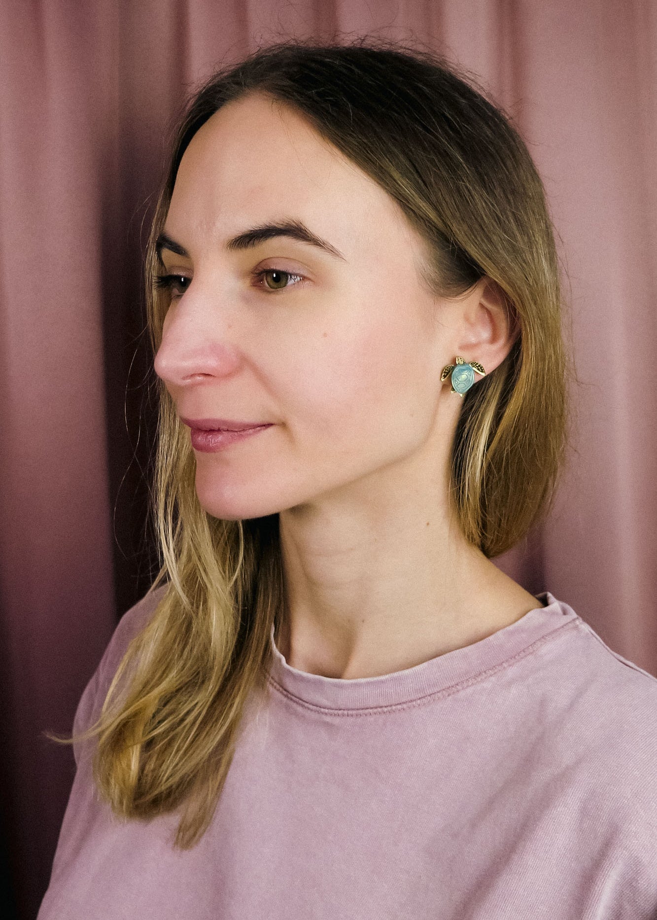 A woman with straight, light brown hair and light skin wears a light pink top and turquoise Sea Turtle Studs, standing in front of a mauve curtain, gazing slightly to the left for a summery vibe.