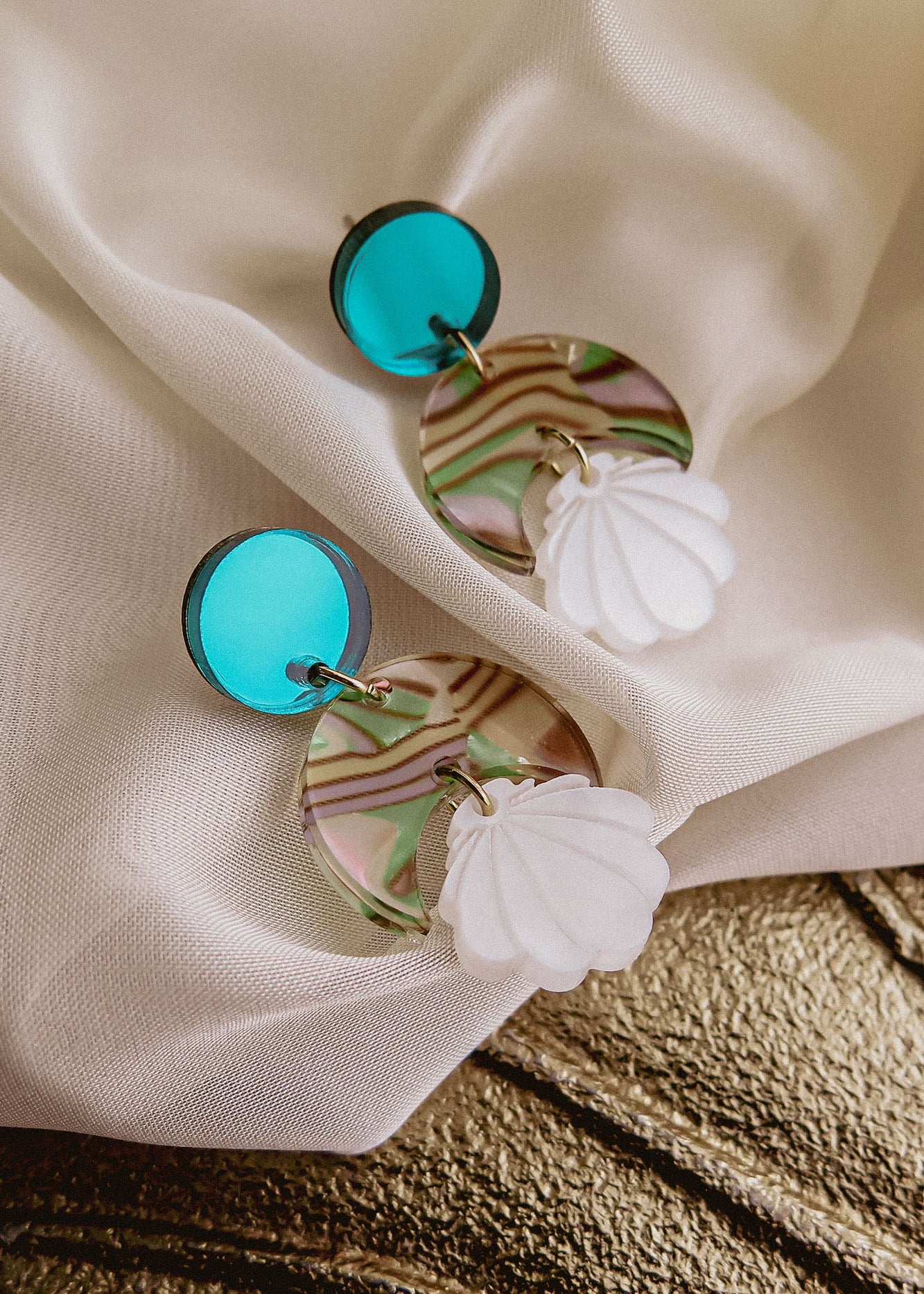 A pair of handmade Seashell Earrings featuring round turquoise studs, marbled circular pieces in green, pink, and clear tones, and lightweight white seashell-shaped charms, displayed on draped white fabric.