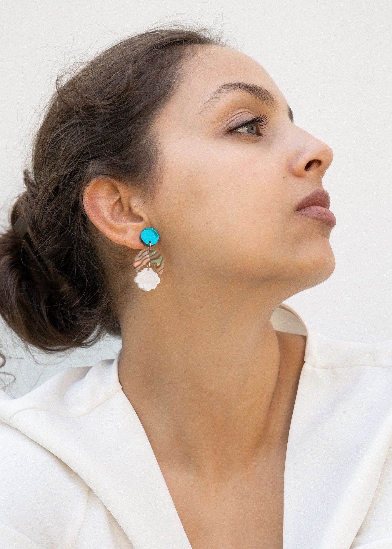 A woman with brown hair in a loose bun wears a white blouse and lightweight statement earrings with turquoise and gold accents. She gazes to the side against a plain, light background.