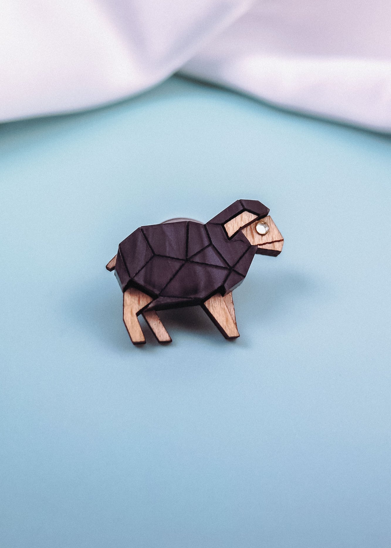A wooden lapel pin shaped like a geometric ram with black and natural wood tones sits on a light blue surface, perfect for animal lovers and fans of polygon design, with white fabric in the background.