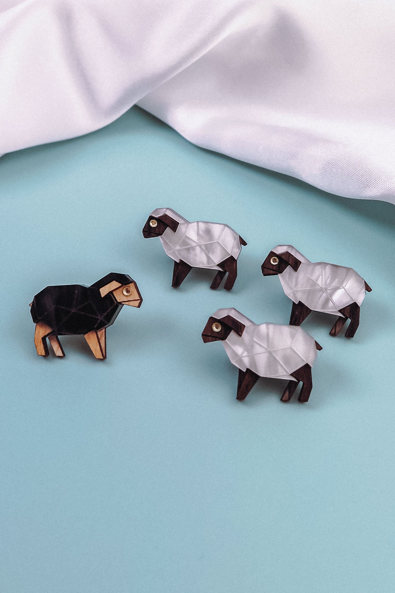 Four geometric sheep figurines on a light blue surface—three are white with brown accents, and one is black with tan accents. A white cloth is in the background, perfect for animal lovers seeking unique decor or a Sheep Badge-inspired accent.