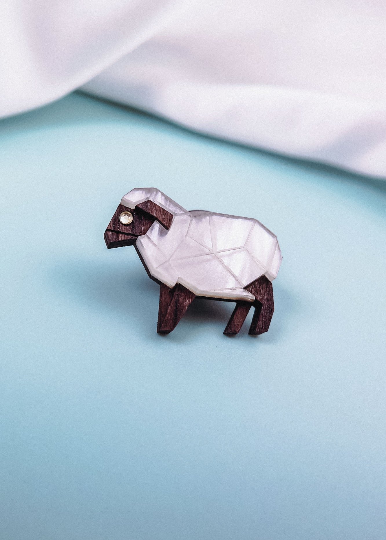 A small, geometric sheep figurine with a white, faceted body and dark brown legs and head, placed on a light blue surface—perfect inspiration for an animal lovers’ Sheep Badge or decorative lapel pin.