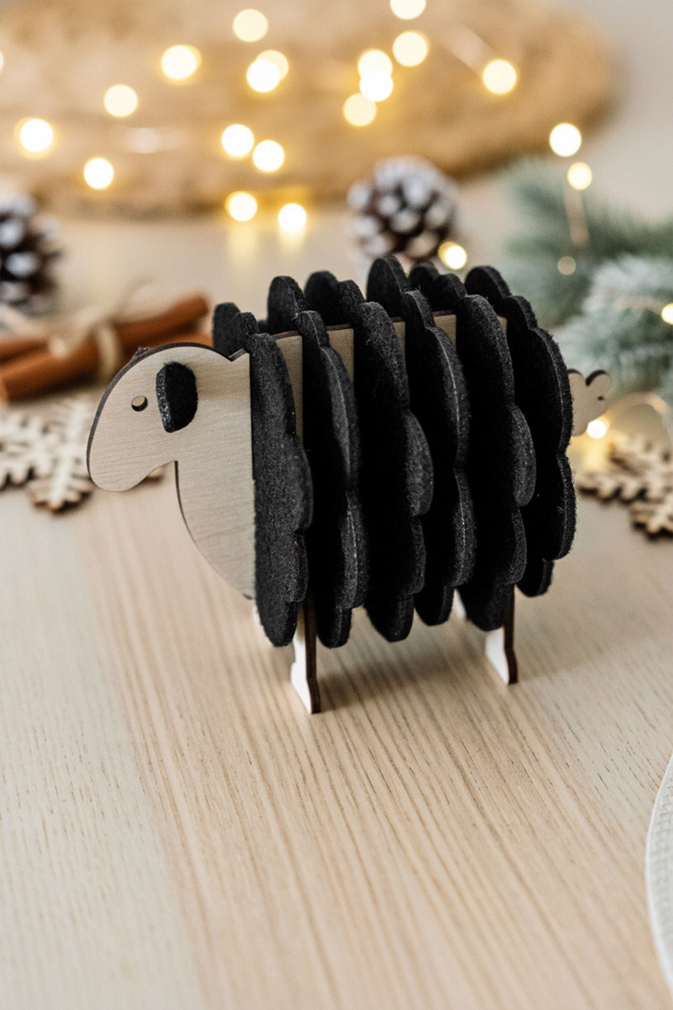 A small wooden sheep figurine made with black felt adds charm to your home decor, standing on a light wooden table with festive lights, pinecones, and greenery blurred in the background.