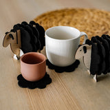 A white mug and a pink cup rest on felt coasters, accompanied by charming wooden sheep coaster holders on a light wooden table. Perfect as home decor accents, with a woven placemat in the background.
