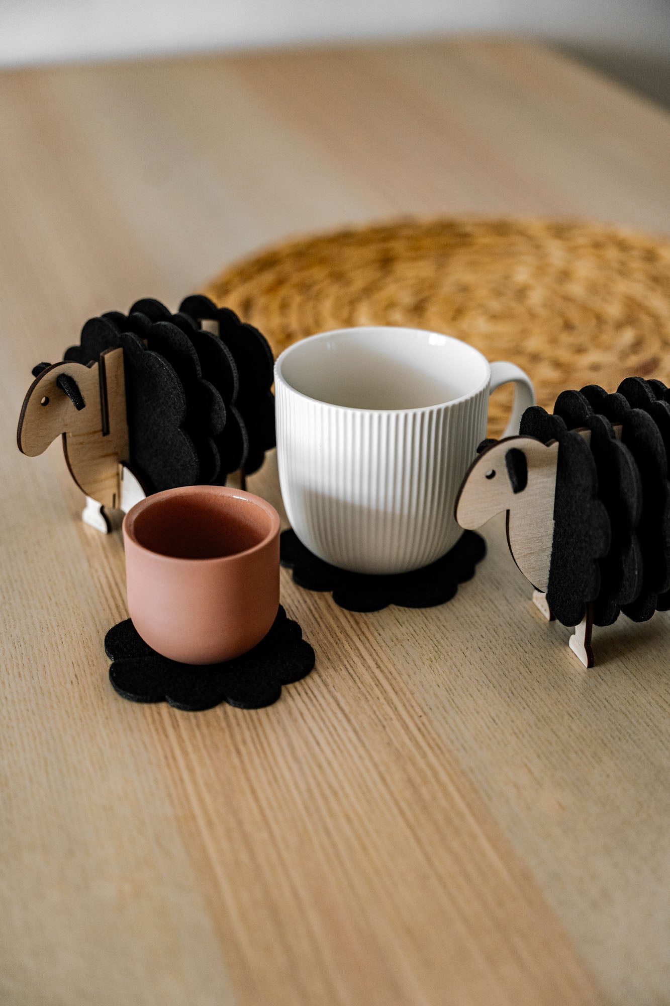 A white mug and a pink cup rest on felt coasters, accompanied by charming wooden sheep coaster holders on a light wooden table. Perfect as home decor accents, with a woven placemat in the background.