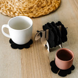 A white mug and a pink cup sit on black felt coasters next to a wooden sheep-shaped coaster holder on a light wooden table with a woven placemat in the background, perfect for charming home decor.
