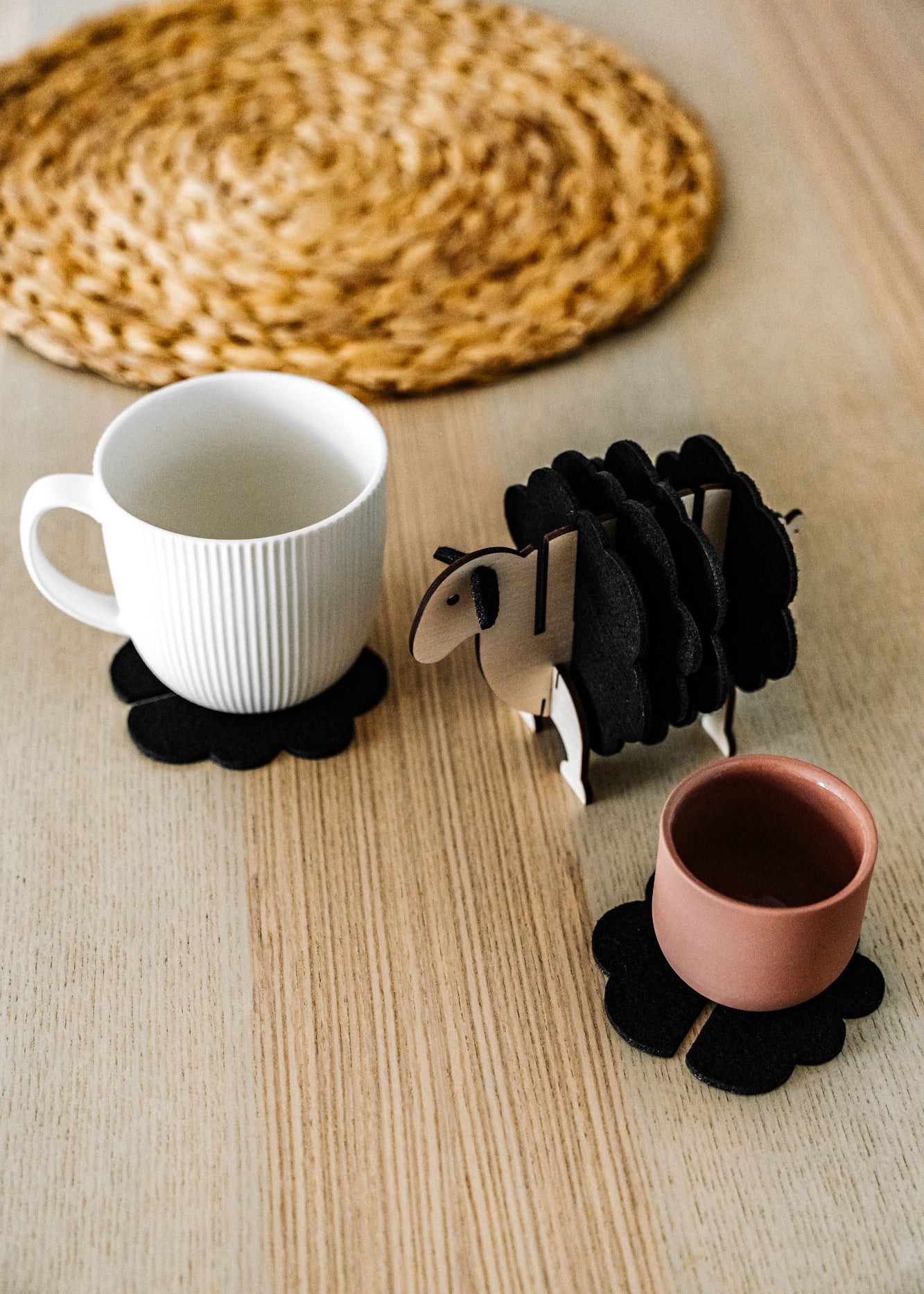 A white mug and a pink cup sit on black felt coasters next to a wooden sheep-shaped coaster holder on a light wooden table with a woven placemat in the background, perfect for charming home decor.