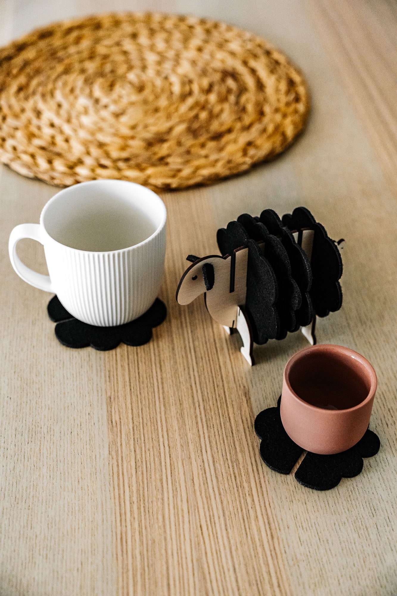 A white mug and a pink cup sit on black felt coasters next to a wooden sheep-shaped coaster holder on a light wooden table with a woven placemat in the background, perfect for charming home decor.