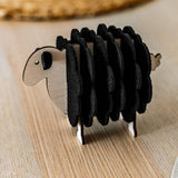 A charming wooden sheep-shaped coaster holder with black felt coasters stands on a light wooden table, bringing a touch of playful home decor alongside a woven placemat and white plate in the background.