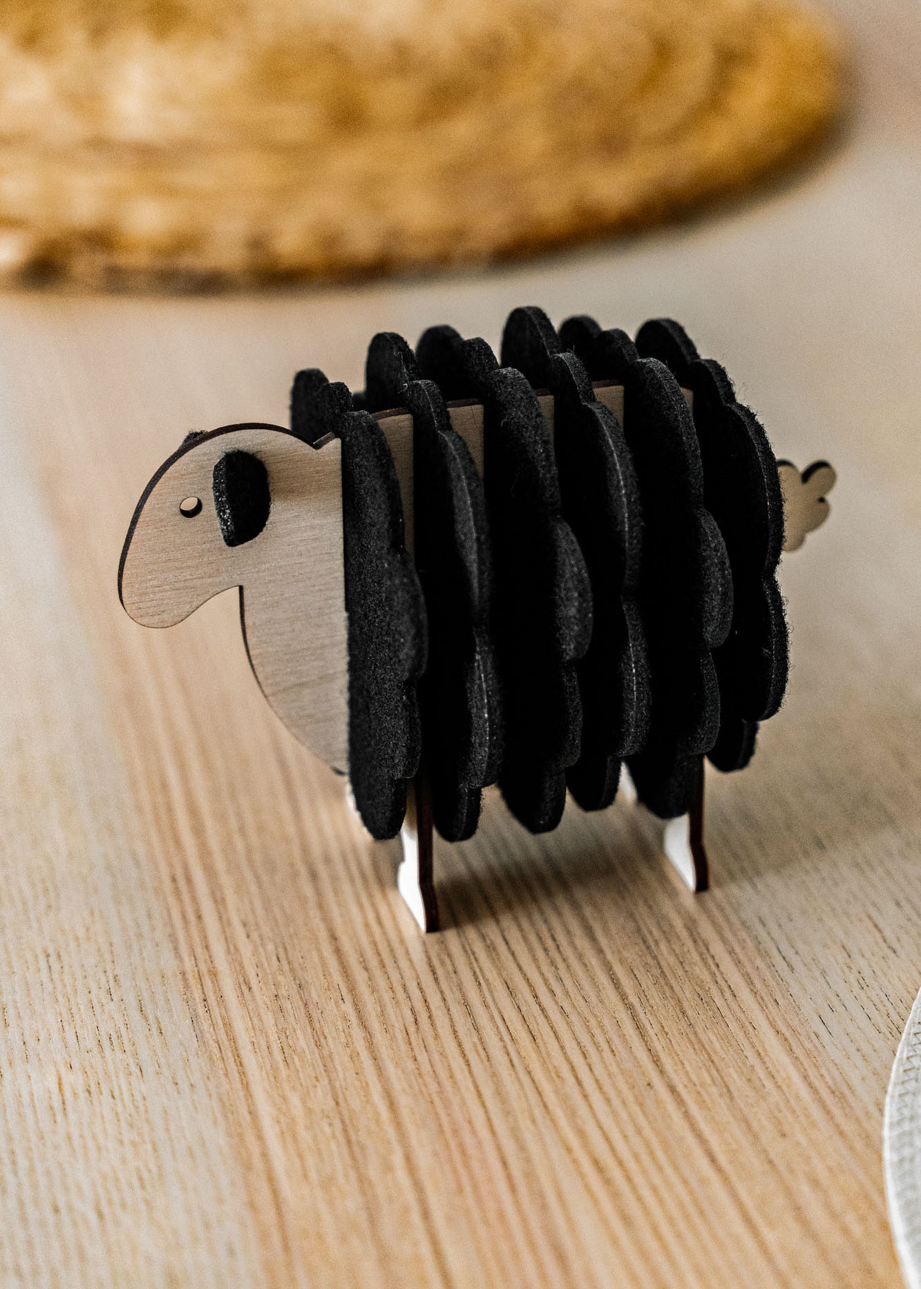 A charming wooden sheep-shaped coaster holder with black felt coasters stands on a light wooden table, bringing a touch of playful home decor alongside a woven placemat and white plate in the background.