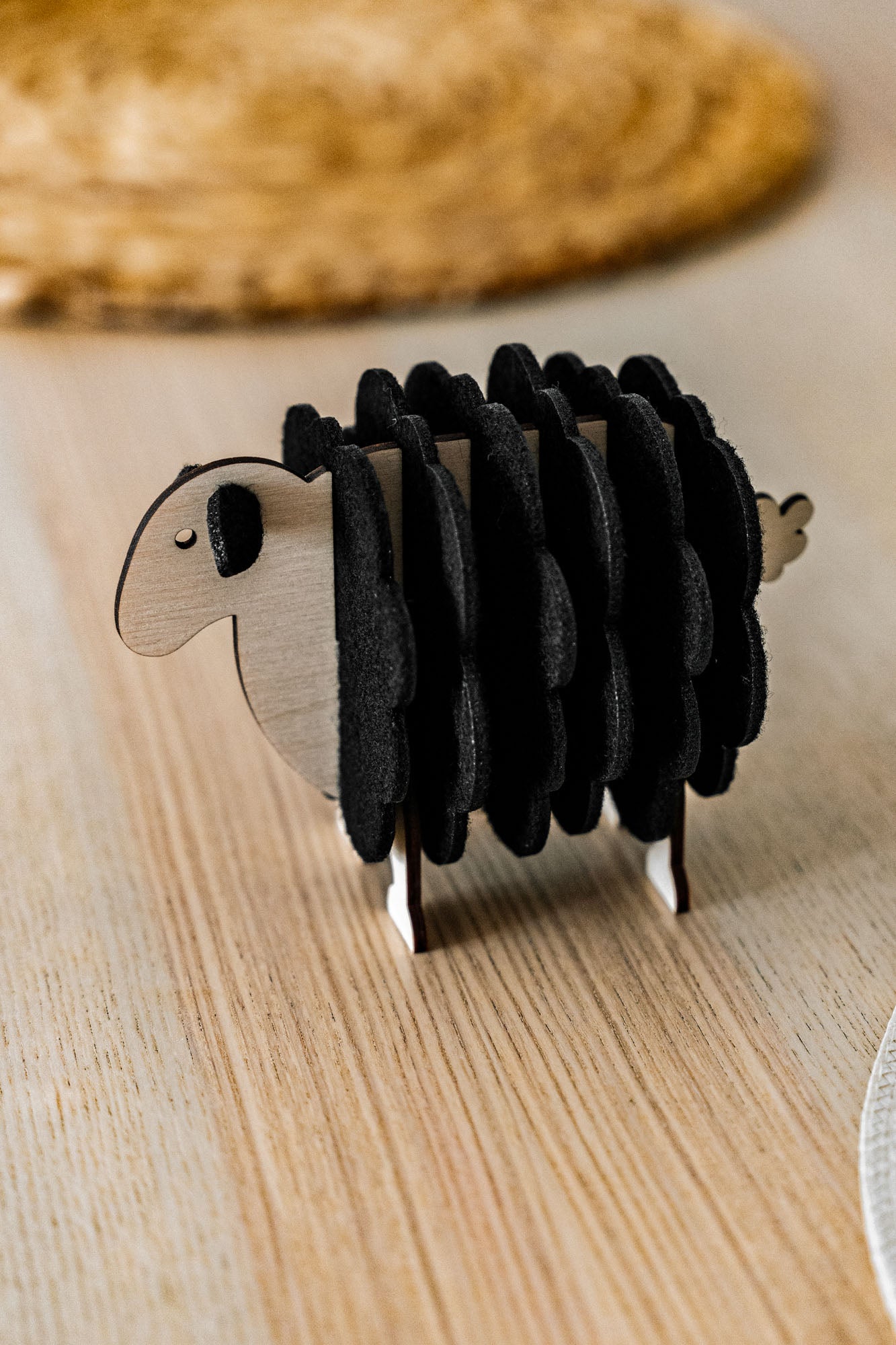 A charming wooden sheep-shaped coaster holder with black felt coasters stands on a light wooden table, bringing a touch of playful home decor alongside a woven placemat and white plate in the background.