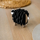 A small, black and white wooden sheep-shaped figure sits on a light wooden table near woven placemats, adding a charming touch to your home decor.
