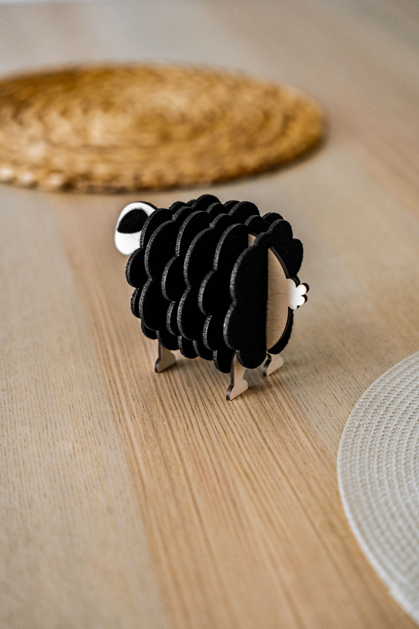 A small, black and white wooden sheep-shaped figure sits on a light wooden table near woven placemats, adding a charming touch to your home decor.
