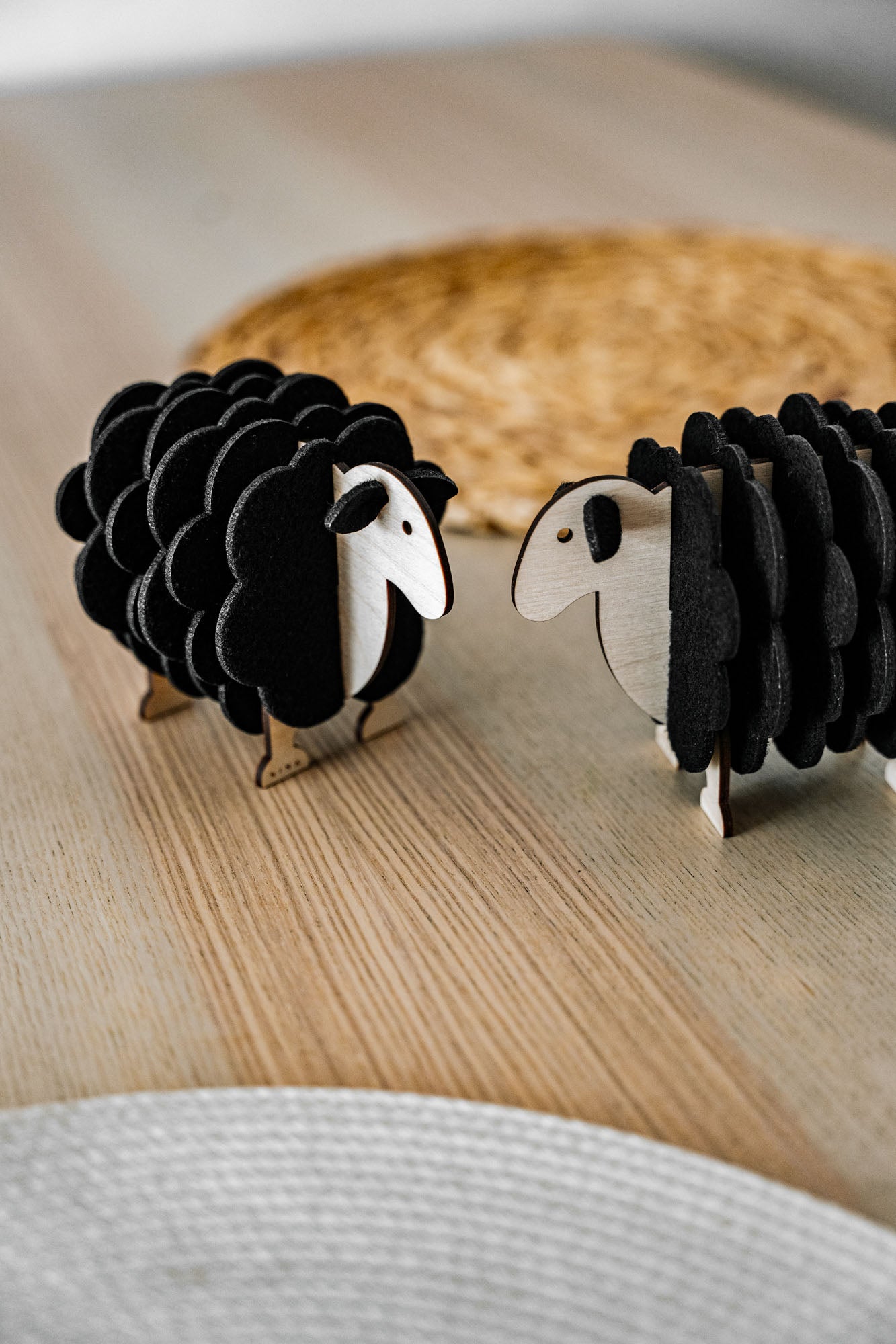 Two small, black wooden sheep figurines made of wood and felt stand facing each other on a light wooden surface, perfect for adding charm to your home decor, with a woven mat blurred in the background.