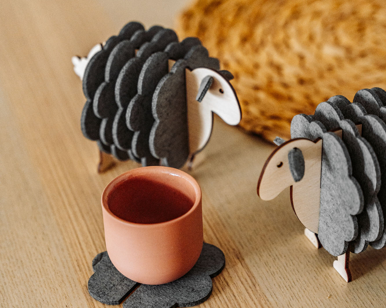 A pink cup rests on a gray, flower-shaped felt coaster beside two wooden sheep figurines—charming home decor—placed on a light wooden surface, with a woven mat partially visible in the background.