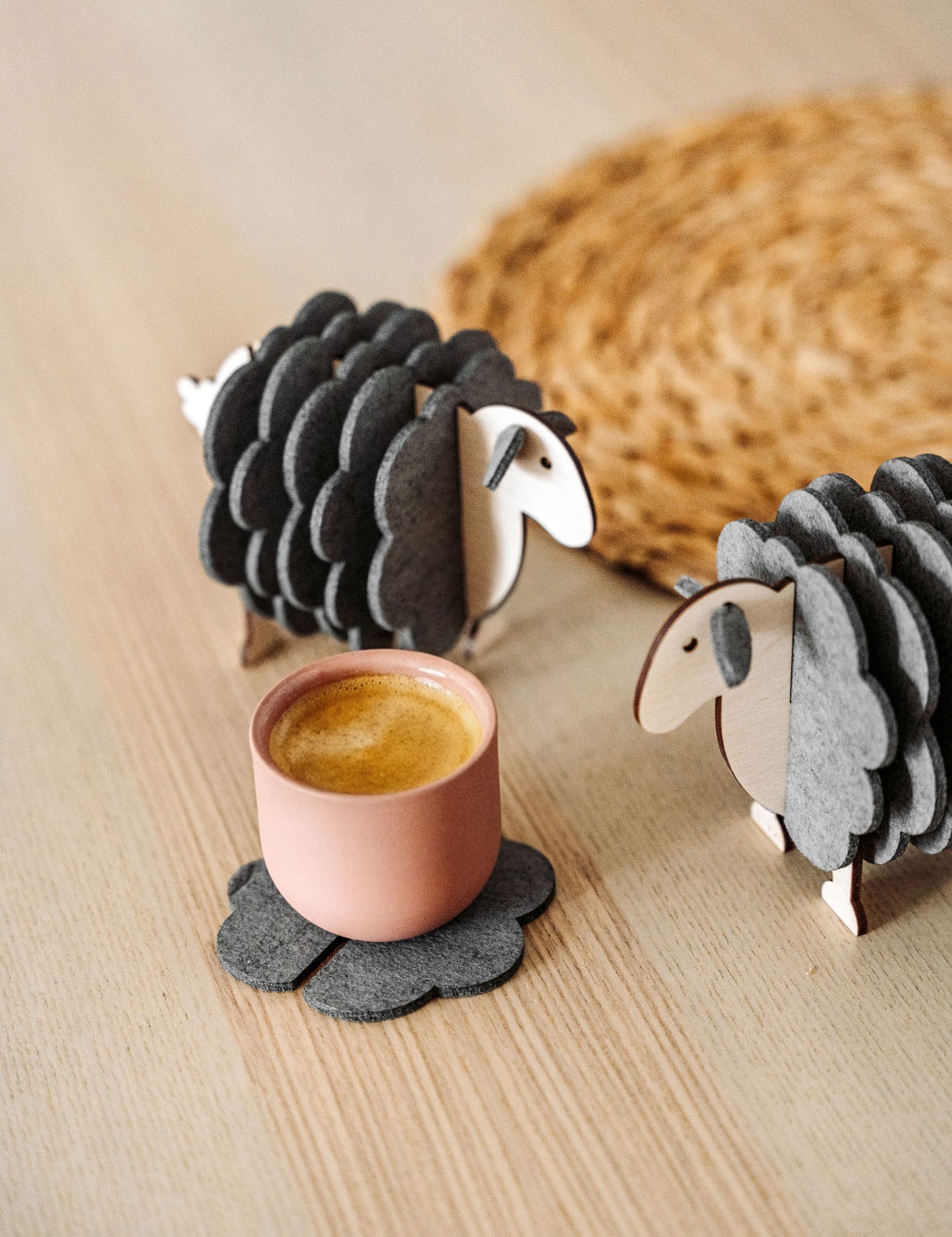 A pink cup rests on a gray, flower-shaped felt coaster beside two wooden sheep figurines—charming home decor—placed on a light wooden surface, with a woven mat partially visible in the background.
