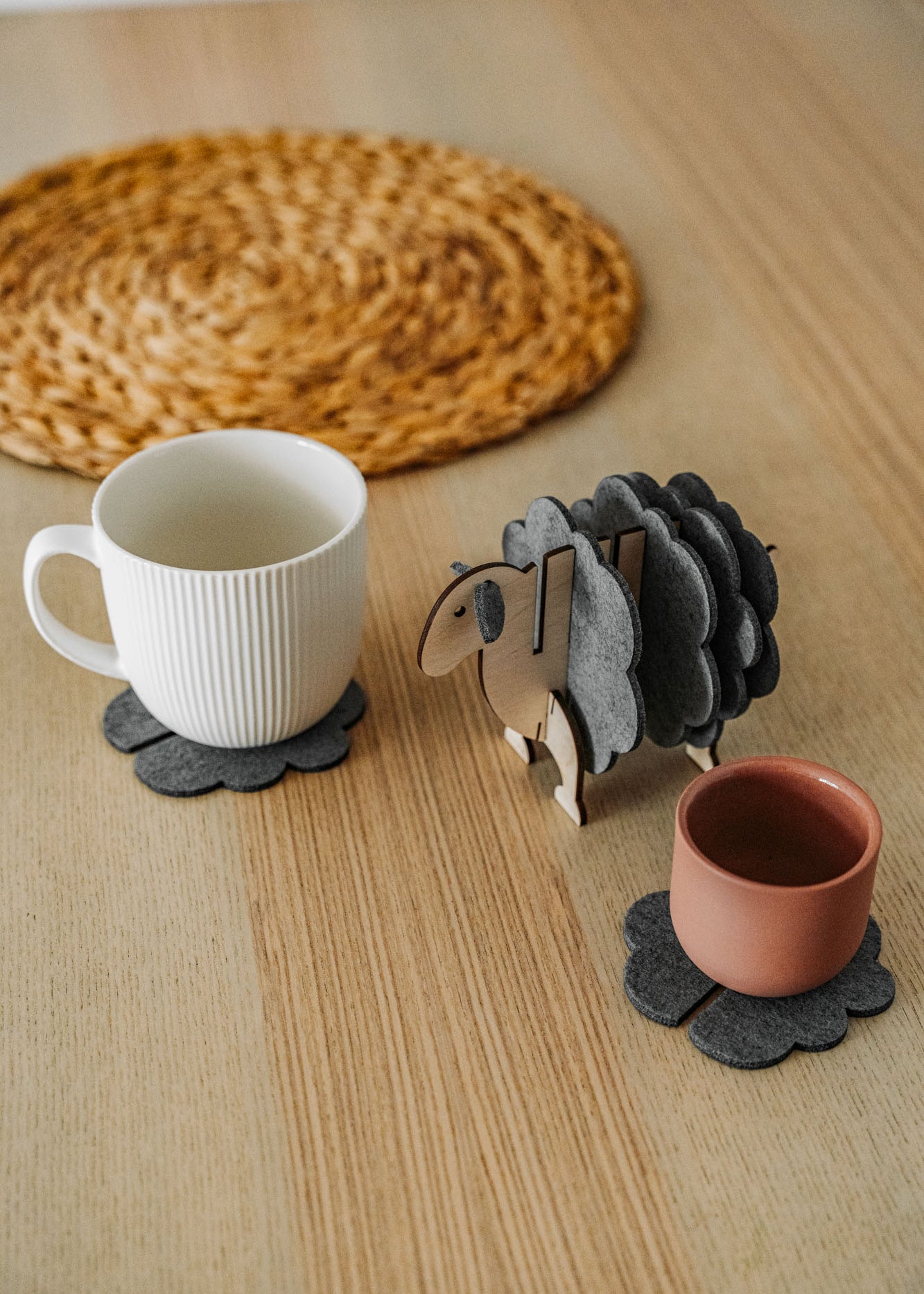 A white mug and a pink cup sit on dark felt coasters next to a decorative wooden sheep-shaped coaster holder, bringing charming home decor touches to a light wooden table topped with a round woven placemat.