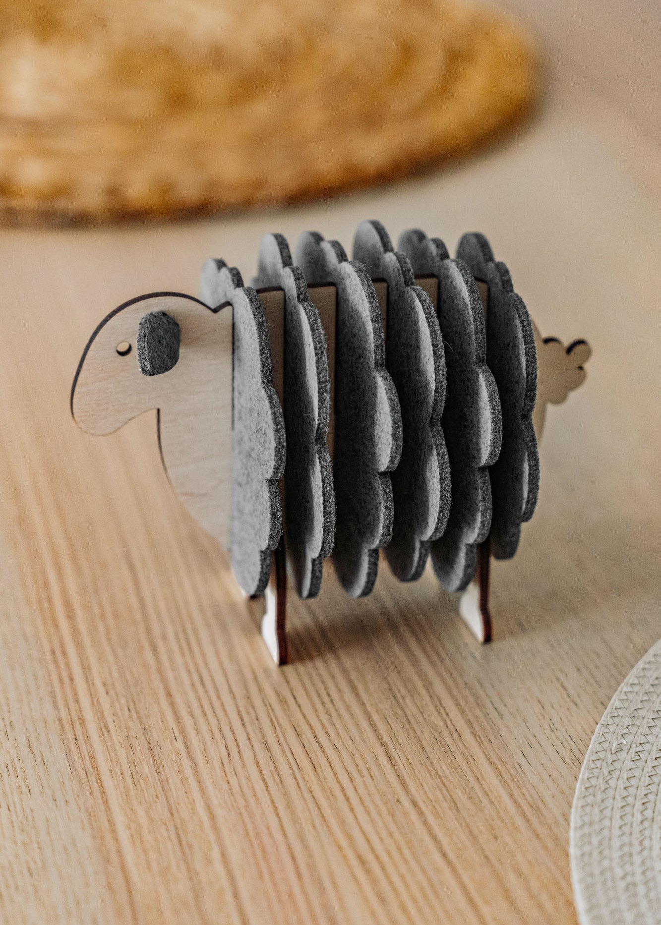 A wooden sheep-shaped coaster holder with round, gray felt coasters arranged to resemble the sheep’s wool stands on a light wooden table—perfect for charming home decor.