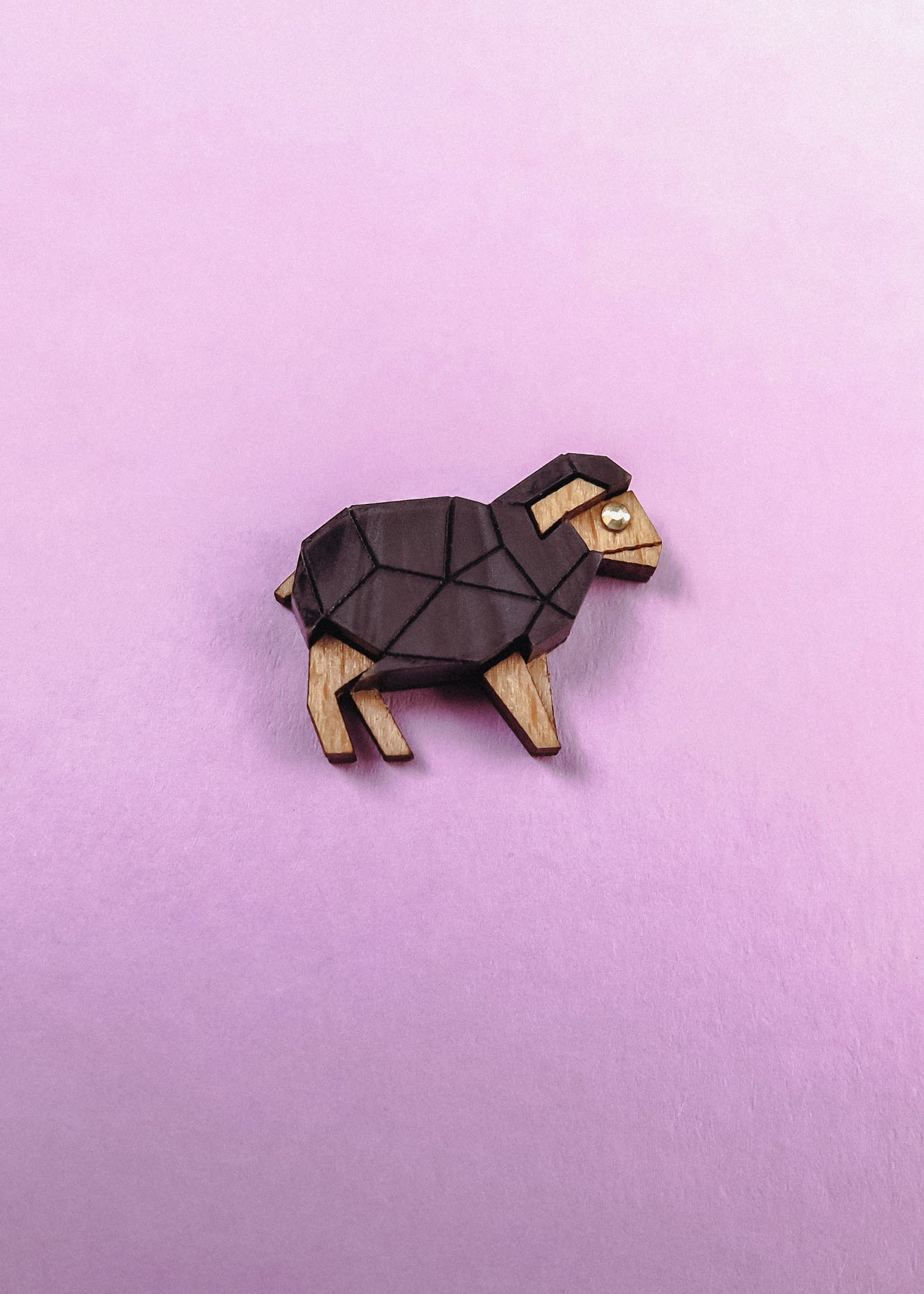 A small, geometric goat figurine with a polygon design, crafted from wood and dark material, sits on a pastel pink background. The minimalist piece features angular shapes—perfect for animal lovers with a flair for modern decor.