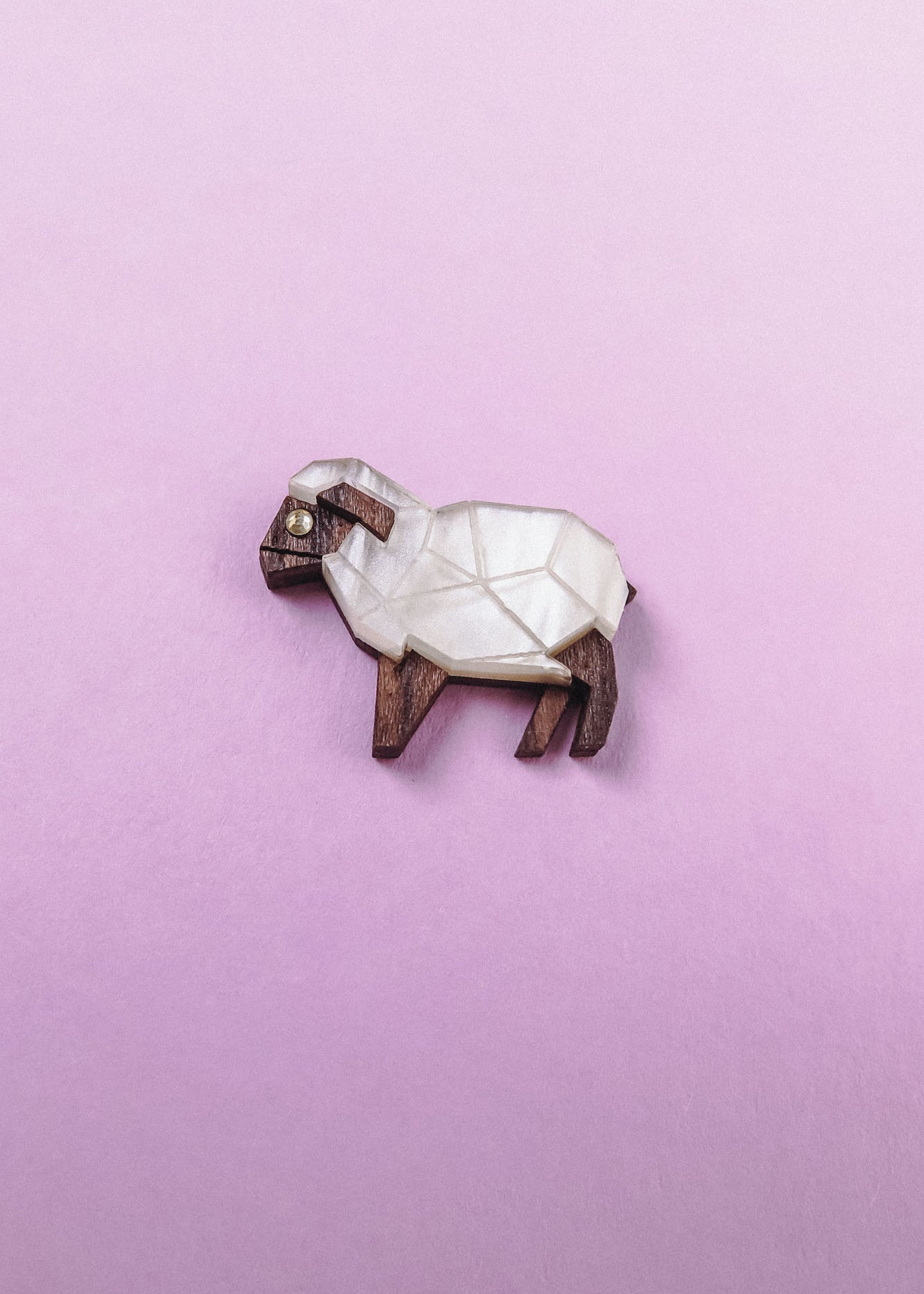 A small, geometric sheep figurine made of wood and white material sits on a pastel pink background—an ideal Sheep Badge or keepsake for animal lovers.