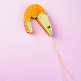 A Shrimp Pin with orange, gold, and green details is placed on a light purple background. Long thin strings extend from the brooch, mimicking shrimp antennae—making it a playful accessory and unique wardrobe charm.